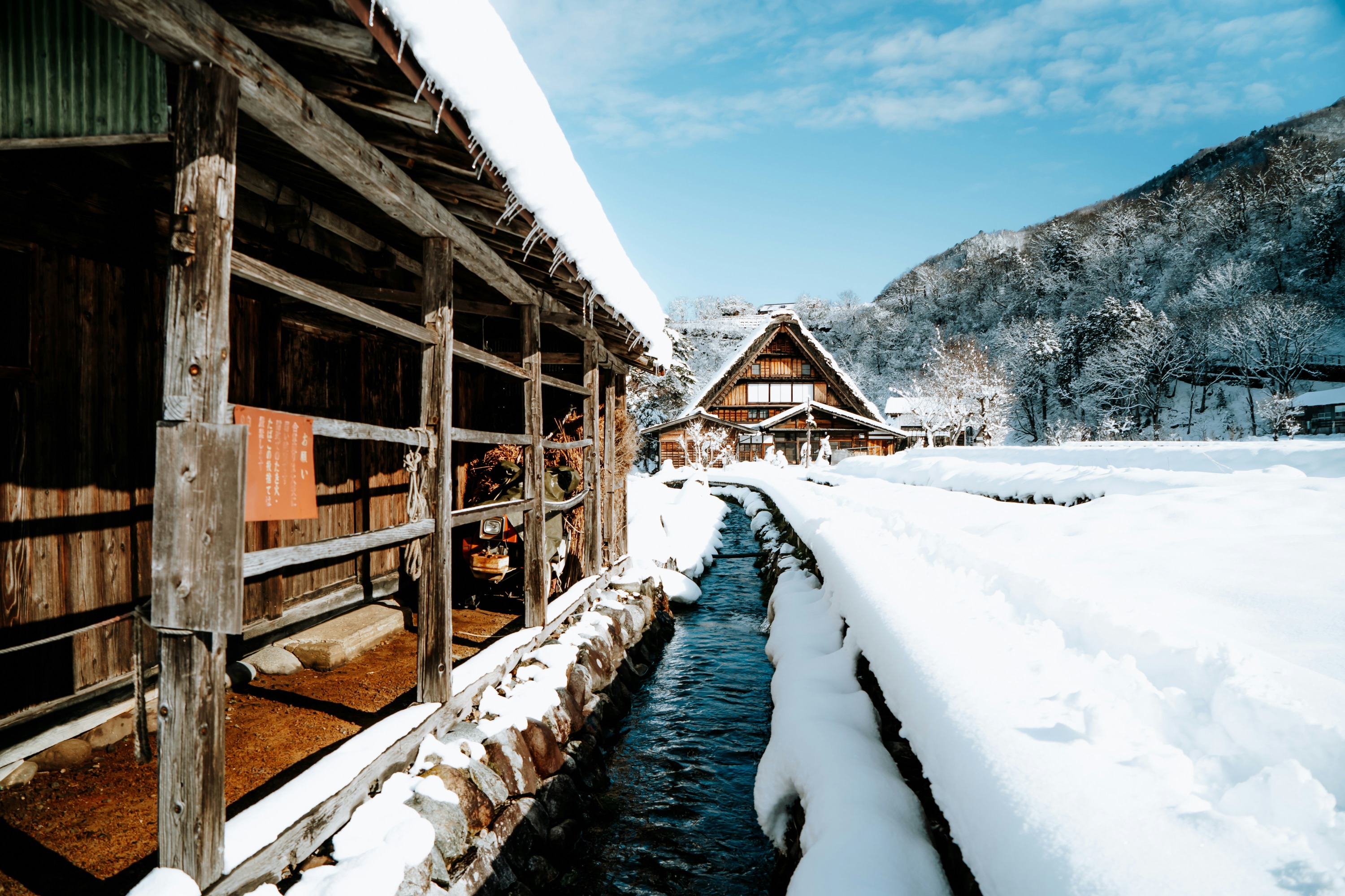 A picturesque winter scene in the historic village of Shirakawa-go, Japan, on a bright, sunny day. In the foreground, a man-made channel with clear, flowing water cuts through the landscape, its stone banks heavily laden with thick, pristine snow. On the left, a rustic wooden structure with open beams provides a close-up textural element.